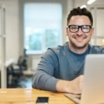 Accueil Happy man wearing glasses working remotely on laptop in modern office environment.